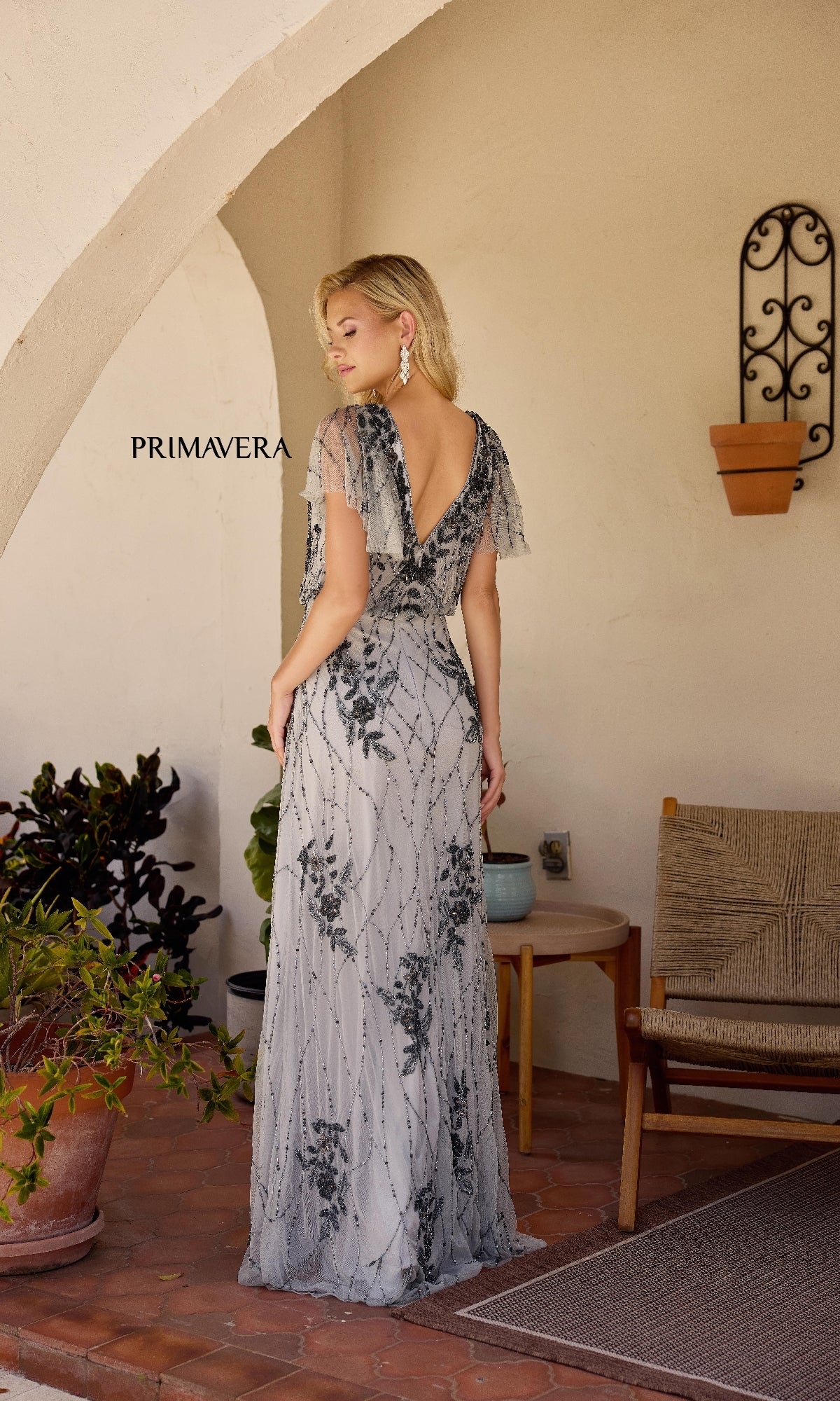 A woman stands on a tiled patio, surrounded by plants and outdoor furniture, wearing the Long Formal Dress: Primavera 13144—a floor-length silver and black beaded gown—with her back to the camera.